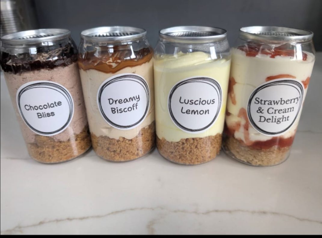 Four Eggless Baker cheesecakes in a can lined up: Chocolate Bliss, Dreamy Biscoff, Luscious Lemon, and Strawberry and Cream Delight